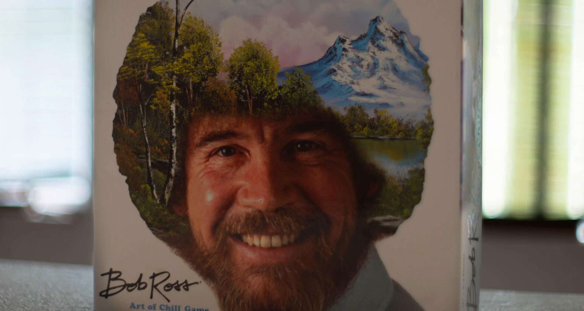 Das Bob Ross-Brettspiel „The Art of Chill“ steht aufrecht auf einem Tisch. Auf dem Deckel ist das Konterfei von Bob Ross zu sehen. Sein lockiges Haar ist mit einer Landschaft mit Bäumen, Bergen, einem See und weißen Wolken am blauen Himmel übermalt.