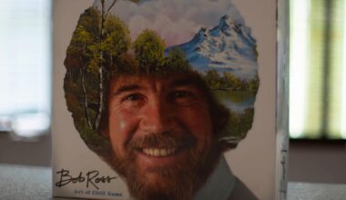 Das Bob Ross-Brettspiel „The Art of Chill“ steht aufrecht auf einem Tisch. Auf dem Deckel ist das Konterfei von Bob Ross zu sehen. Sein lockiges Haar ist mit einer Landschaft mit Bäumen, Bergen, einem See und weißen Wolken am blauen Himmel übermalt.