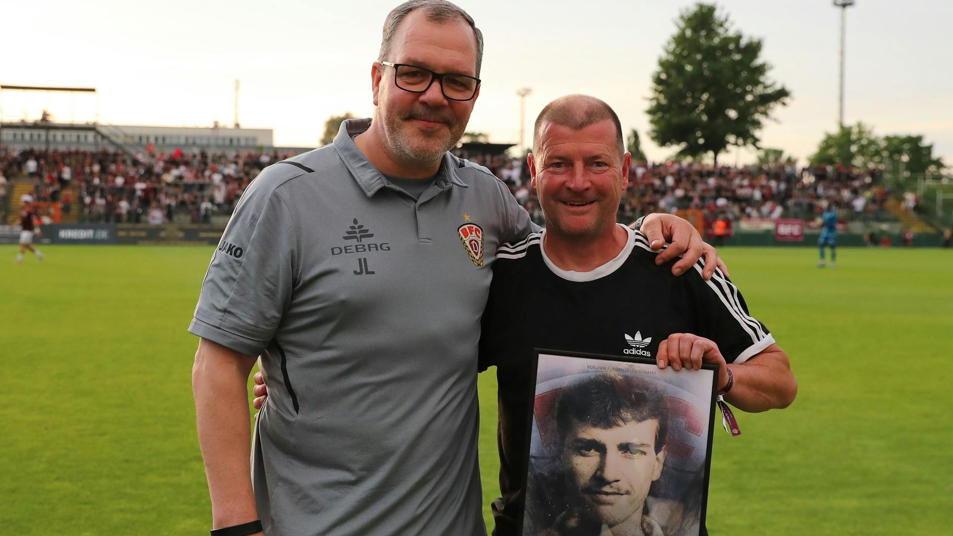 Jens-Uwe Zöphel (r.) hält ein Foto seiner aktiven Zeit in der Hand, lächelt mit Jörn Lenz, Teammanager des BFC Dynamo, in die Kamera.