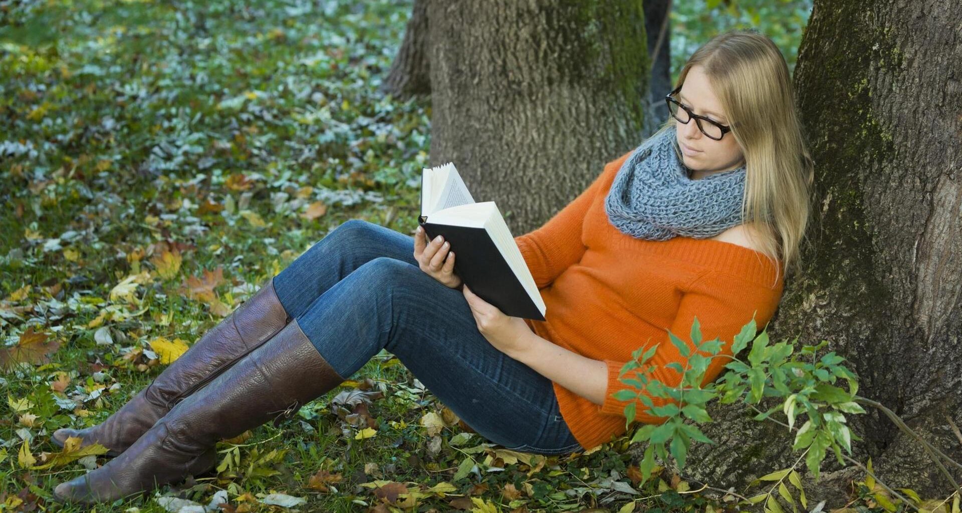 Junge Frau mit Brille sitzt im Park und liest ein Buch.