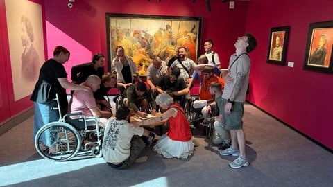 Eine Gruppe von 16 Personen steht mittig im Raum, einige Männer sind im Rollstuhl. Drei Menschen mal ein Bild. An den Wänden hängen Gemälde.