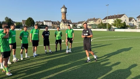 Ganz rechts ist Trainer Julian Feit zu sehen. Dieser hält auf dem Platz des Stadions am Wasserturm in Neuwied-Engers eine Ansprache an seine Spieler. Eingige von ihnen sind links vom Trainer im Bild zu sehen. Im Hintergrund ist das Wahrzeichen von dem Ort Neuwied-Engers zu sehen, der Wasserturm.