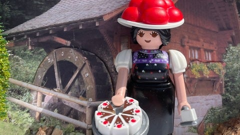 Die Playmobilfigur "Schwarzwald Marie" mit Bollenhut und Torte vor einer kleinen ortstypischen Mühle