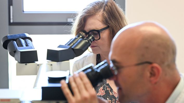 Ina Czyborra (SPD, hinten), Gesundheitssenatorin von Berlin, schaut mit Lars Bullinger, Klinikdirektor Hämatologie, Onkologie und Tumorimmunologie, durch ein Mikroskop auf dem Universitätsmedizin Berlin Campus Virchow-Klinikum am 21.08.2025. (Quelle: picture alliance/dpa/Jens Kalaene)