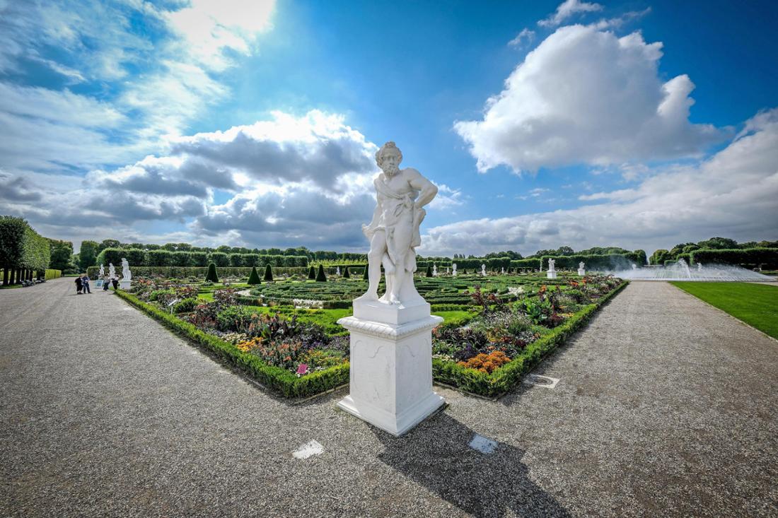 Die Herrenhäuser Gärten in Hannover werden 350 Jahre alt. Das Bild zeigt den Großen Garten, im Vordergrund eine Statue, dahinter Blumenbeete, Wege mit einigen Spaziergängern und ein Springbrunnen sowie angrenzende Hecken.