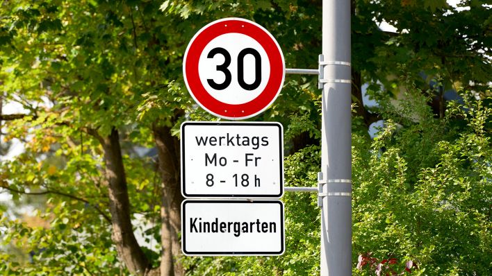 Ein Verkehrsschild in einer grünen Umgebung, das eine Geschwindigkeitsbegrenzung von 30 km/h anzeigt, gültig an Werktagen in der Nähe eines Kindergartens. (Quelle: dpa/Michael Bihlmayer)