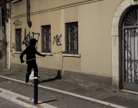 Eine nicht identifizierbare Person mit schwarzem Kapuzenpulli und dunkler Kleidung übermalt in der Nacht Graffiti an einer Hauswand. 