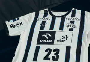 Ein weißes Sporttrikot mit vertikalen schwarzen Streifen, der Nummer 23 und verschiedenen Sponsorenlogos, darunter Puma, Orlen, Star, HAX und Chefs Culinar, sowie dem Vereinsabzeichen des THW Kiel.