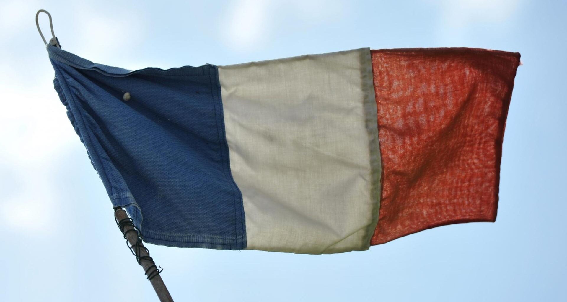 Eine französische Flagge flattert im Wind, dahinter sieht man blauen Himmel mit hellen Wolken