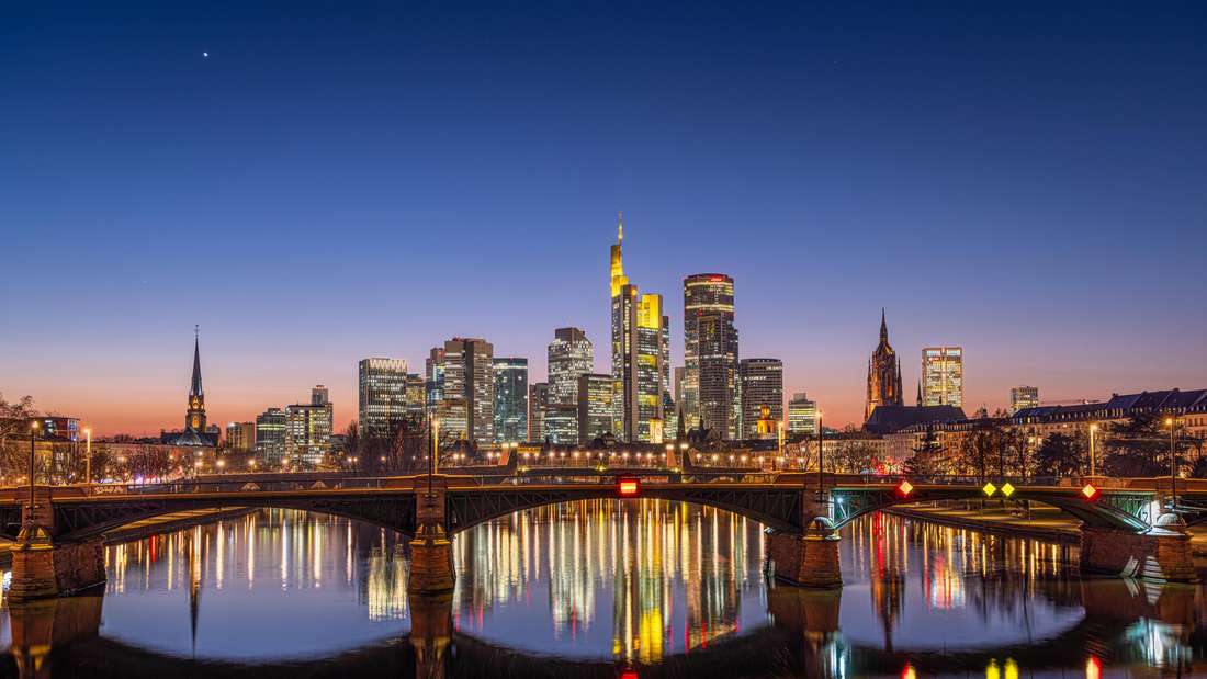 Die Frankfurter Skyline bei Nacht, im Hintergrund einer Brücke über einem Fluss, auf dem sich die Lichter spiegeln.