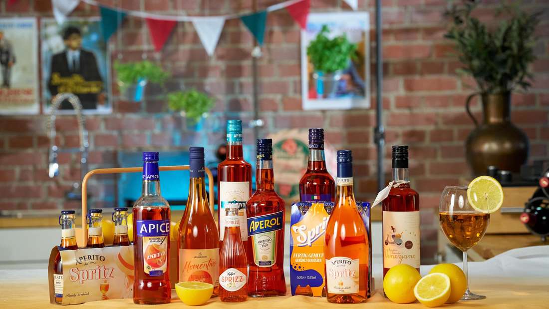Auf einem Tisch für eine Party stehen verschiedene große und kleine Flaschen von Aperol Spritz sowie Gläser und Zitronen aufgereiht.