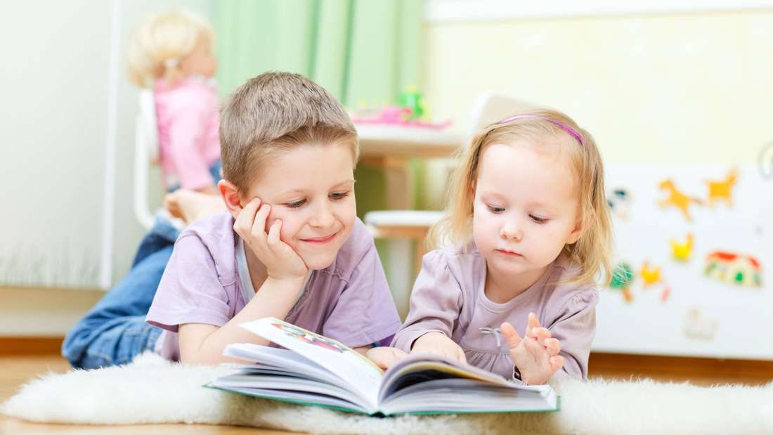 Bruder und Schwester liegen im Kinderzimmer auf dem Boden und lesen zusammen ein Buch (Symbolbild)