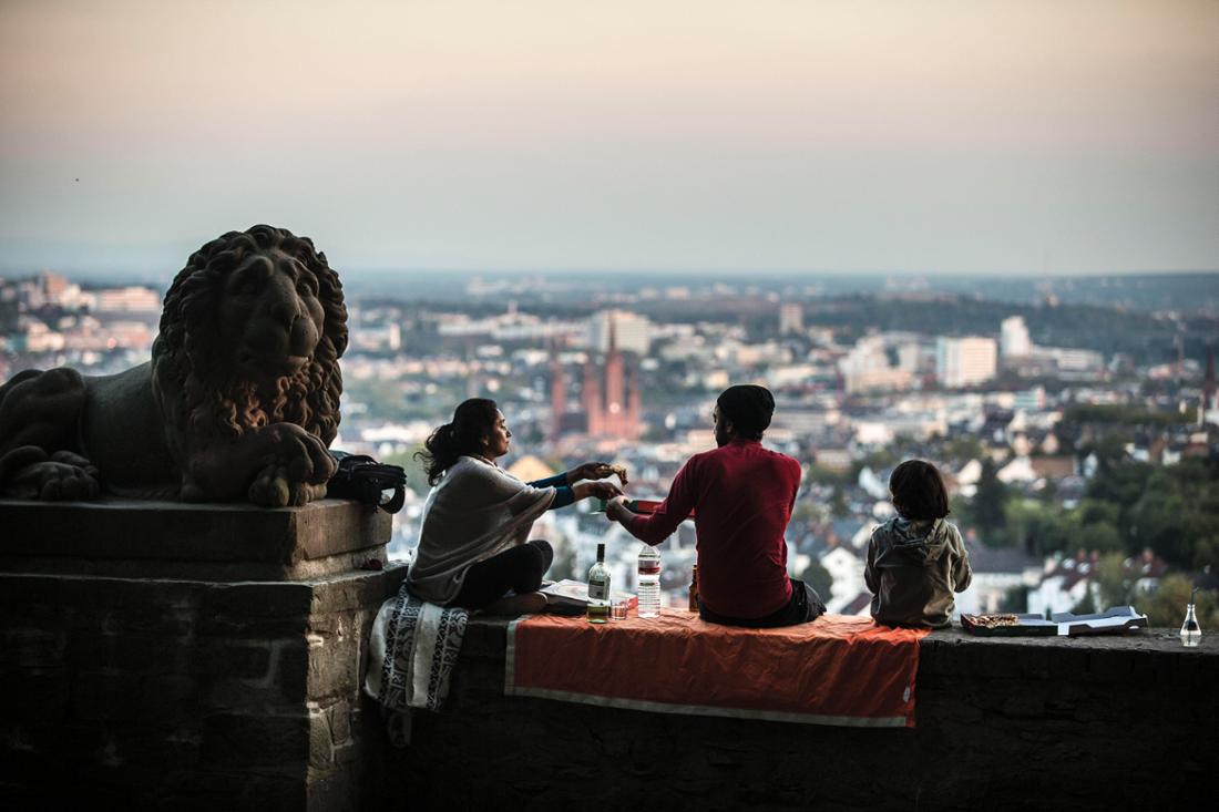 Ein Mann, eine Frau und ein Kind sitzen auf dem Löwenplatz auf dem Neroberg in Wiesbaden und genießen den Ausblick auf die Stadt. Im Hintergrund ist die Marktkirche als roter Backsteinbau zu erkennen. 