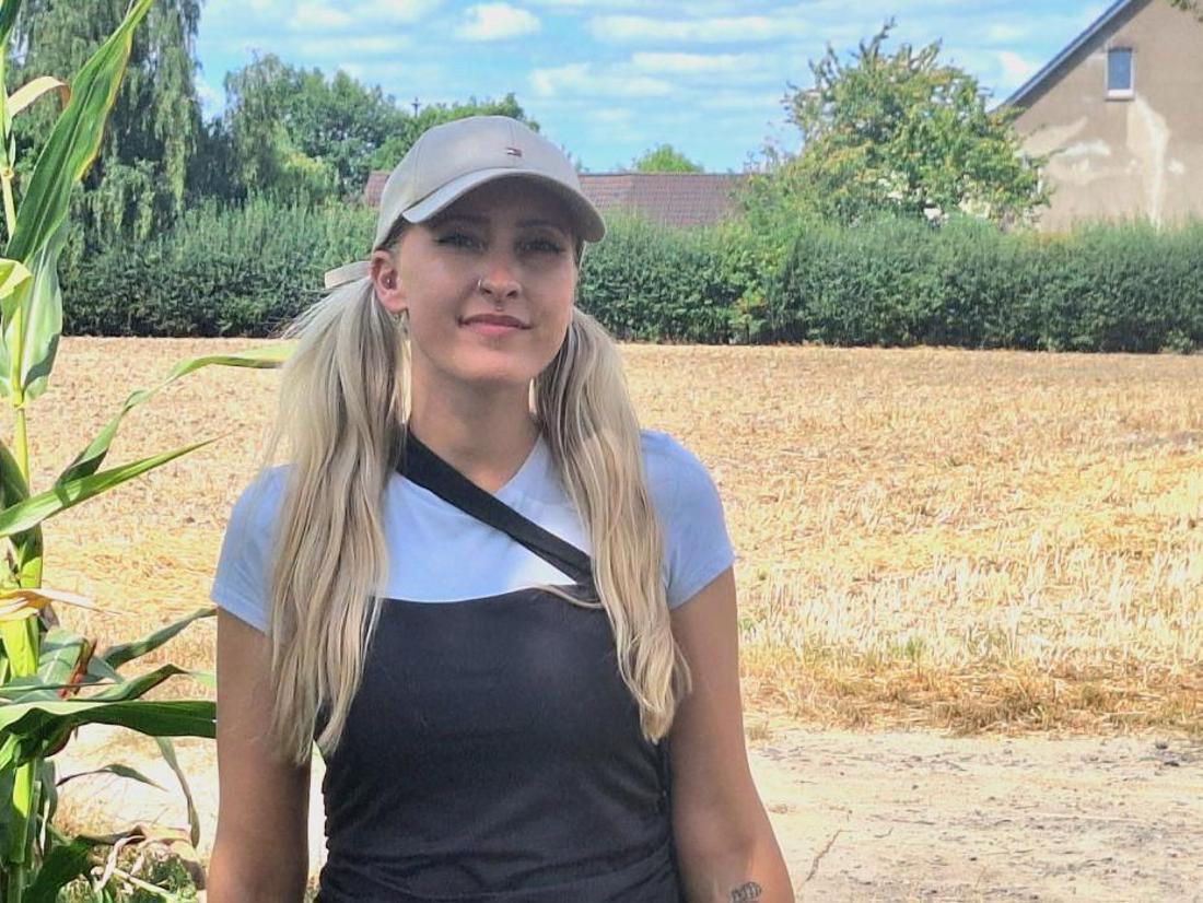 Sarah-Alice ist auf dem Bild zu sehen. Sie steht alleine vor einem Feld und lächelt direkt in die Kamera. 