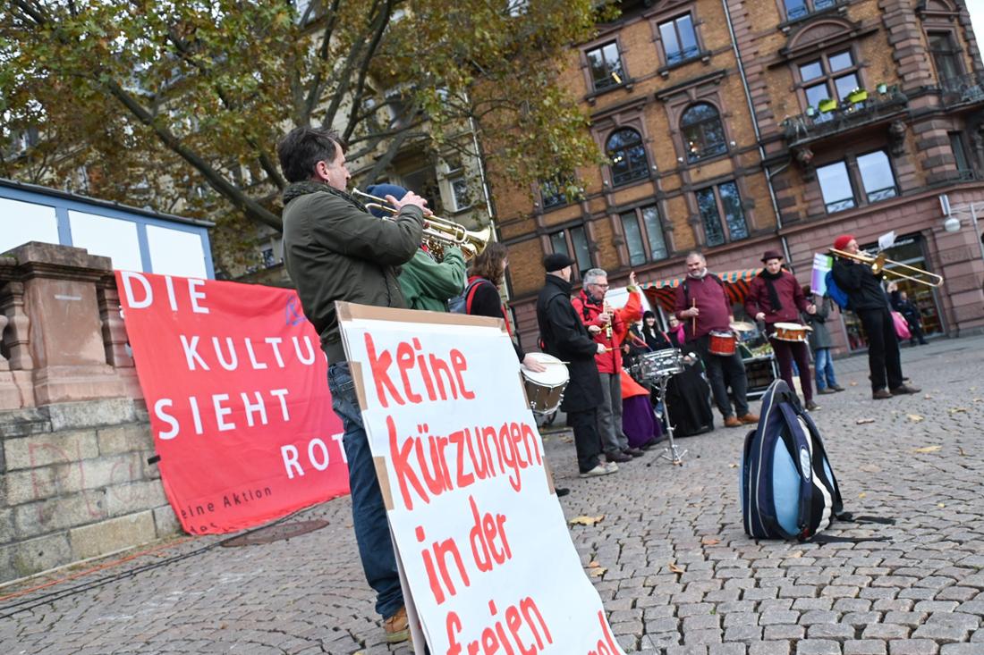 Musiker:innen der Kooperative New Jazz aus Wiesbaden protestieren auf dem Dern‘schen Gelände im Wortsinne mit Pauken und Trompeten gegen Kürzungen des Kulturetats. An einer Mauer hinter ihnen ist ein rotes Plakat angebracht, auf dem mit weißer Schrift geschrieben steht: „Die Kultur sieht rot“. 