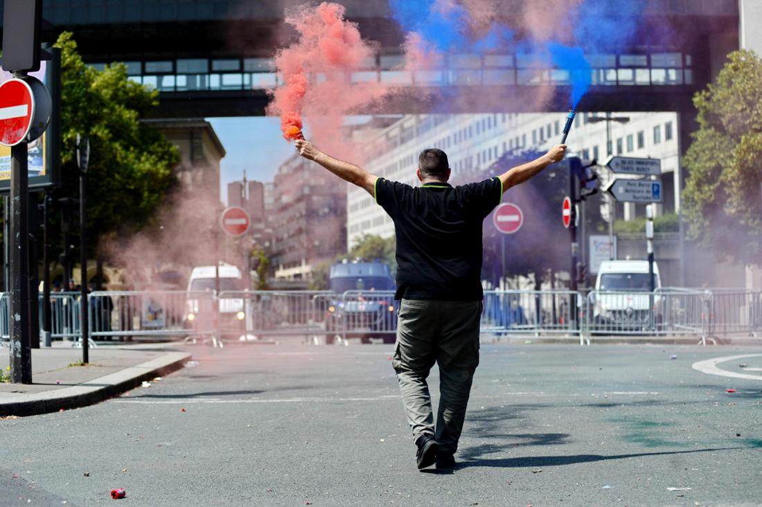 Ein Demonstrant marschiert auf eine Blockade gegen Fahrzeuge zu, er hält dabei zwei Rauchfackeln mit blauem und rotem Rauch in die Höhe. 