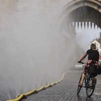 Abkühlung an heißen Tagen: Eine Radfahrerin fährt an einer Wassersprühanlage in Köln vorbei.