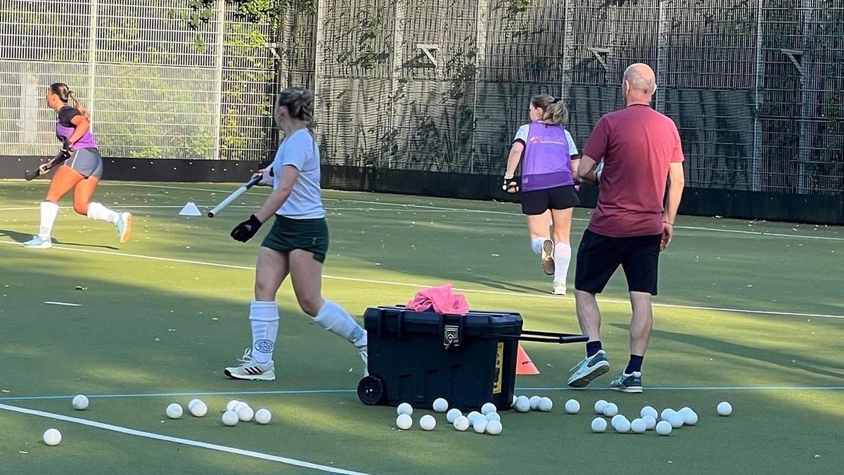 Training bei den Hockeydamen vom HC Essen: Drei Spielerinnen rennen mit Schläger über den Platz. Ihr Trainer steht auf dem Feld und beobachtet. Im Vordergrund liegen viele Hockeybälle.