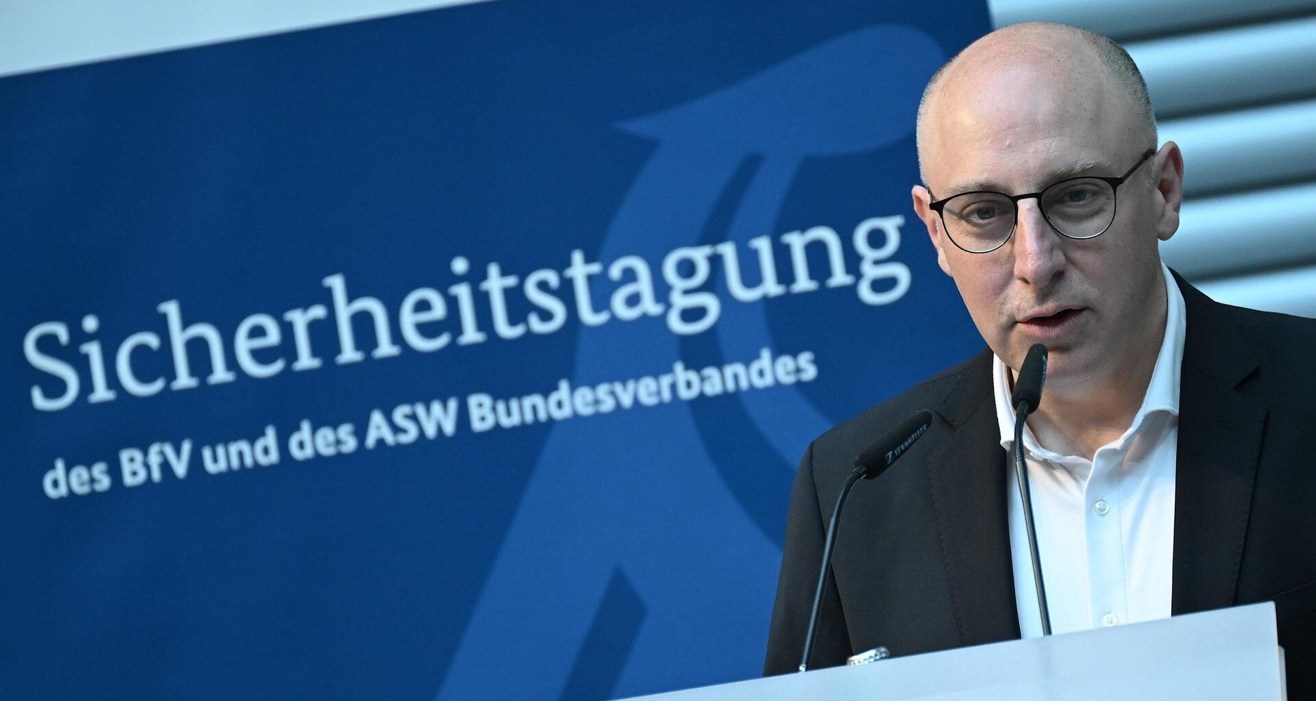 Sinan Selen, Vizepräsident des Bundesamtes für Verfassungsschutz spricht bei einer Sicherheitstagung. Er steht vor einem Mikrofon. Im Hintergrund sieht man ein blaues Plakat mit dem Titel der Veranstaltung.