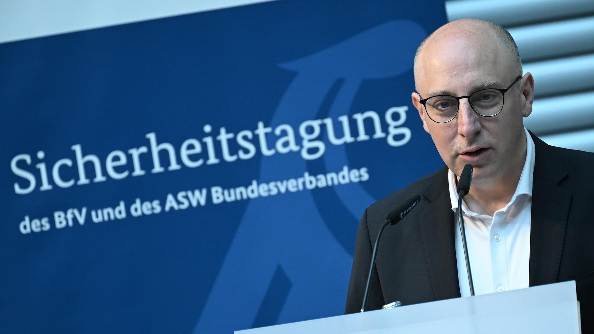 Sinan Selen, Vizepräsident des Bundesamtes für Verfassungsschutz spricht bei einer Sicherheitstagung. Er steht vor einem Mikrofon. Im Hintergrund sieht man ein blaues Plakat mit dem Titel der Veranstaltung.