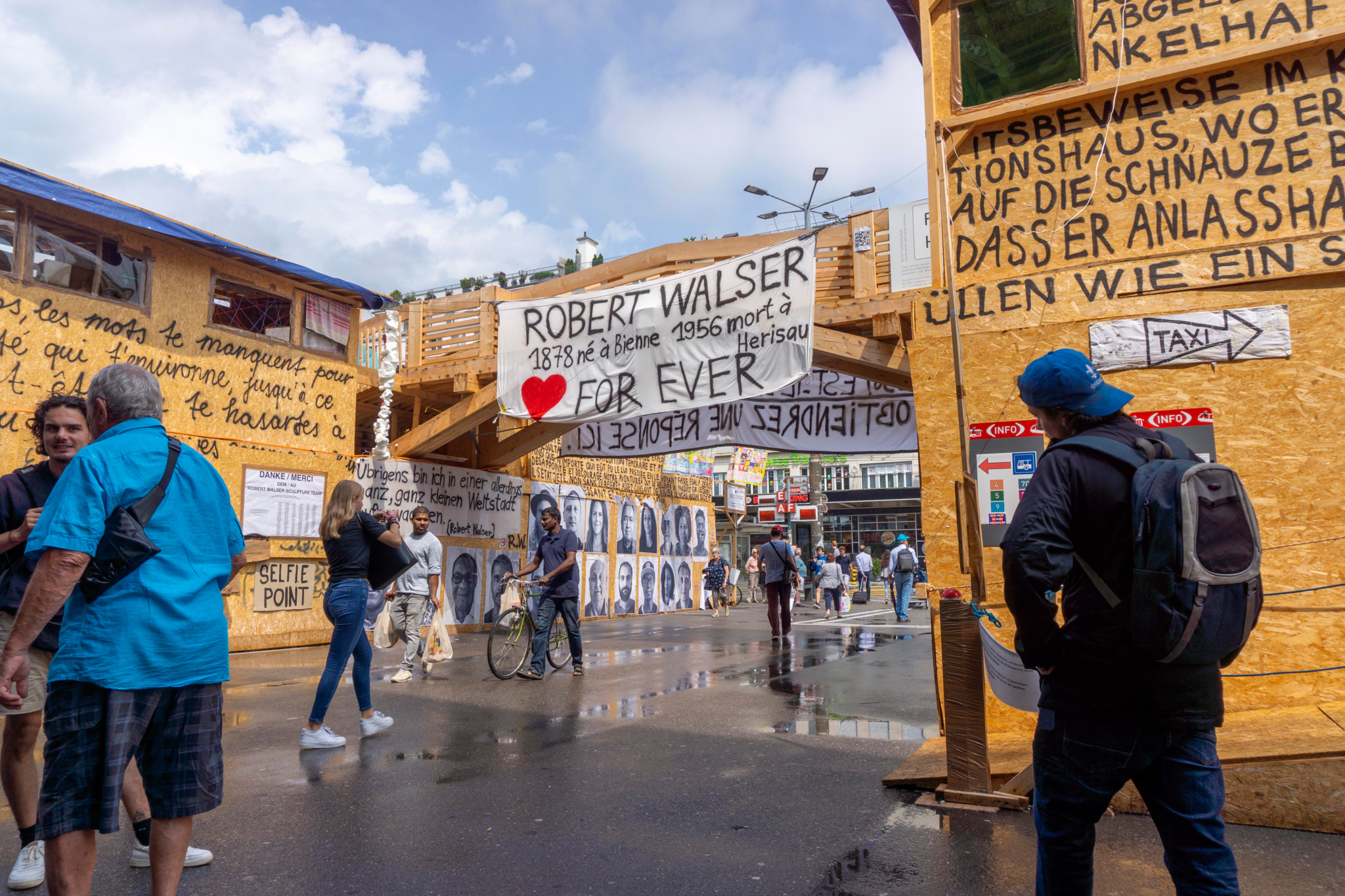 Öffentlicher Ausstellungsraum in Biel, gestaltet von Thomas Hirschhorn, mit Fokus auf Robert Walser. Menschen bewegen sich im kunstreich mit Texten und Porträts dekorierten Bereich.