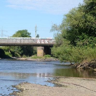 Eine Brücke über einen kleinen Fluss. Der Wasserstand ist niedrig, das Kiesufer ist breit.