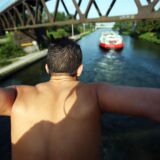 Ein Mann steht mit ausgebreiteten Armen auf einer Brücke über einem Kanal. Im Hintergrund ist ein Frachtschiff zu sehen, das auf dem Wasser fährt.
