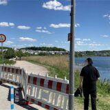Ein Fußgänger steht am Ufer des Phoenix-Sees in Dortmund-Hörde auf einem Weg, der durch rot-weiße Absperrungen teilweise blockiert ist. Ein Schild deutet darauf hin, dass der Weg für Fußgänger und Fahrräder geeignet ist.