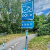 Das Bild zeigt links Heide Kröger-Brenner vom ADFC Dortmund vor einem Abschnitt des Emscherweges. Rechts ist der Emscherweg zu sehen, gekennzeichnet mit einem blauen Schild der Emschergenossenschaft.