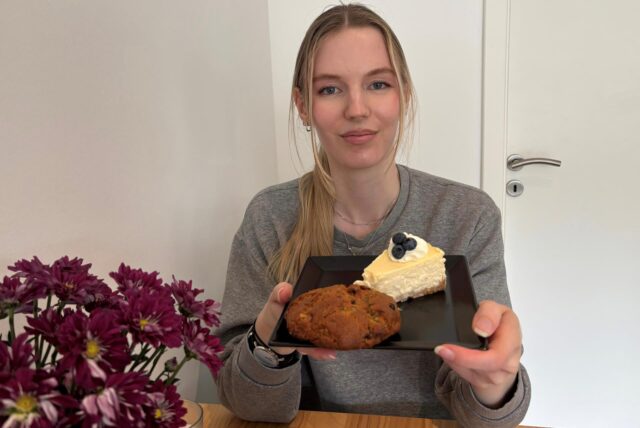 Eine junge Frau präsentiert auf einem schwarzen Teller ein Stück Käsekuchen, garniert mit Blaubeeren und Sahne, sowie einen Keks. Im Vordergrund ist ein Strauß roter Chrysanthemen zu sehen.