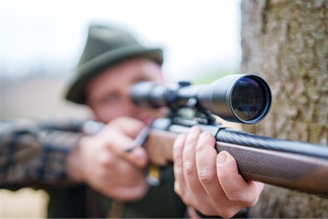 Ein Mann mit Hut zielt mit einem Gewehr, das mit einem Zielfernrohr ausgestattet ist. Der Schütze befindet sich im Freien, möglicherweise während der Jagd oder beim Sportschießen.
