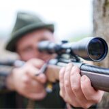 Ein Mann mit Hut zielt mit einem Gewehr, das mit einem Zielfernrohr ausgestattet ist. Der Schütze befindet sich im Freien, möglicherweise während der Jagd oder beim Sportschießen.
