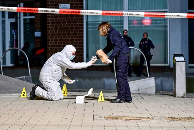 Ein Polizeibeamter im weißen Ganzkörperanzug kniet hinter einer Polizeiabsperrung und reicht einem Polizeibeamten in Uniform etwas.