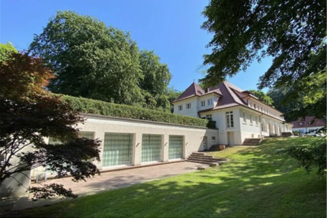 Großes denkmalgeschütztes Anwesen mit weißer Fassade, roten Dachziegeln, mehreren Etagen, großen Fenstern und einer weitläufigen, parkähnlichen Gartenanlage mit altem Baumbestand.