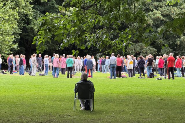 Rund 100 Menschen stehen im Kreis auf einer Wiese im Stadtgarten in Recklinghausen. Eine Frau im Klappstuhl sitzt am Rand und schaut zu.