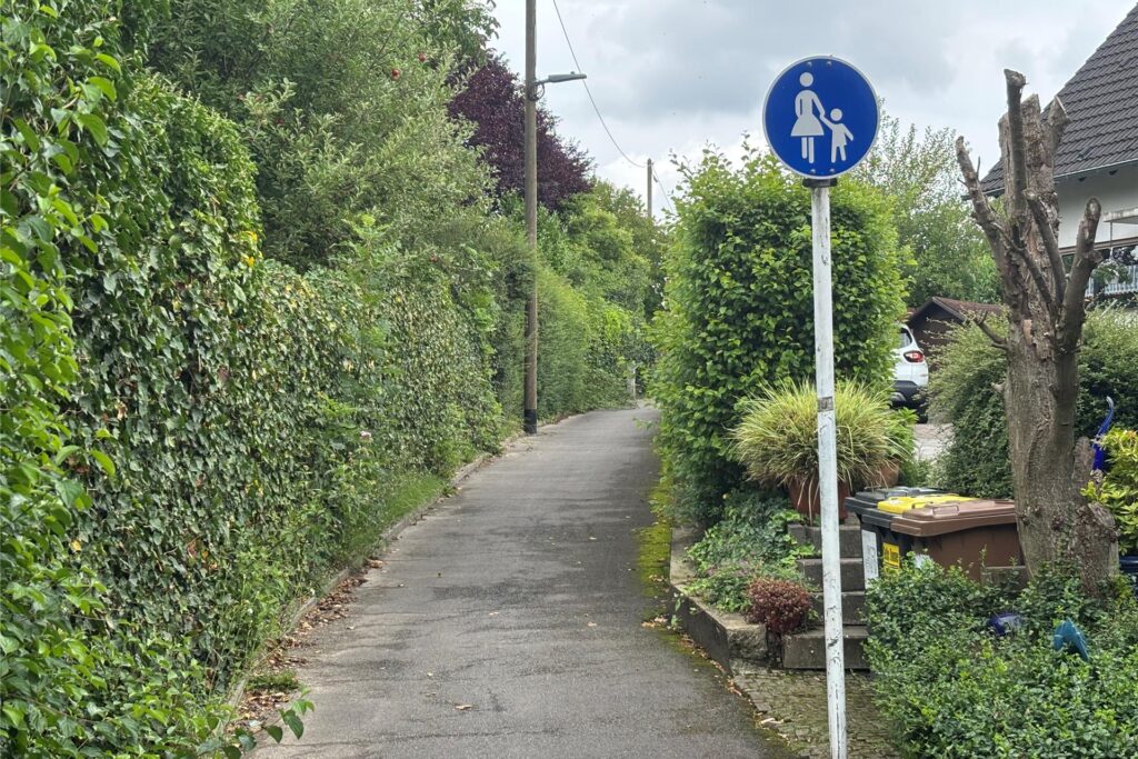 Ein Gehweg ist von üppigem Grün umgeben. Ein blaues Verkehrsschild, das eine Mutter mit Kind darstellt, ist im Vordergrund zu sehen.