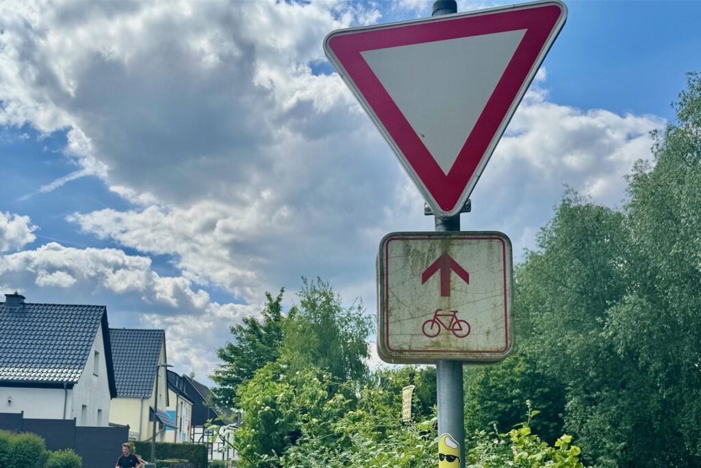 Aufnahme einer Wohnstraße mit Verkehrsschildern, die auf einen bevorrechtigten Fahrradweg hinweisen. Im Hintergrund sind Passanten und Wohnhäuser zu sehen.