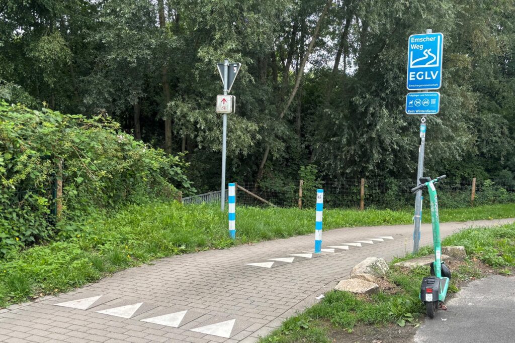 Ein Geh- und Radweg wird durch blaue Markierungen begrenzt. Am Wegesrand ist ein E-Scooter abgestellt.