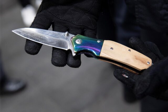 Hände mit schwarzen Handschuhen halten ein Messer in die Kamera.