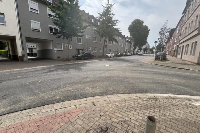 Eine Straßenansicht zeigt Wohnhäuser entlang einer Straßenecke, mit geparkten Autos am Straßenrand. Der Himmel ist bewölkt.
