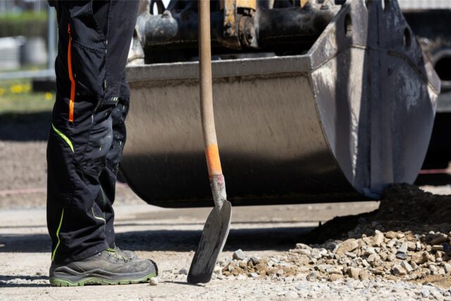 Bauarbeiter mit einer Schaufel vor ienem Bagger.
