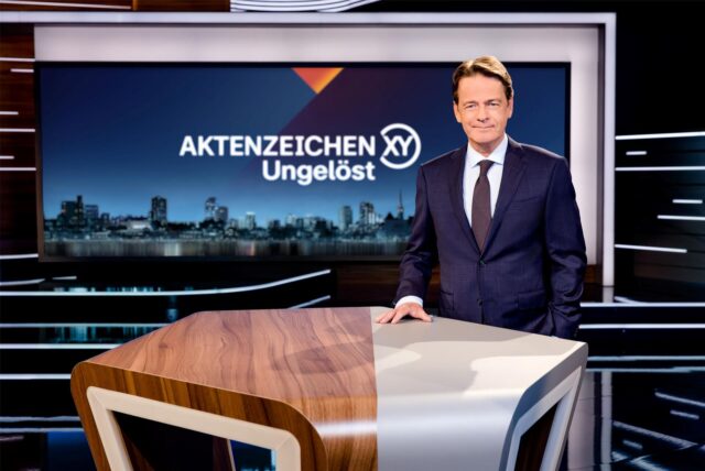 „Aktenzeichen XY… ungelöst“-Moderator Rudi Cerne steht im Studio und lächelt in die Kamera