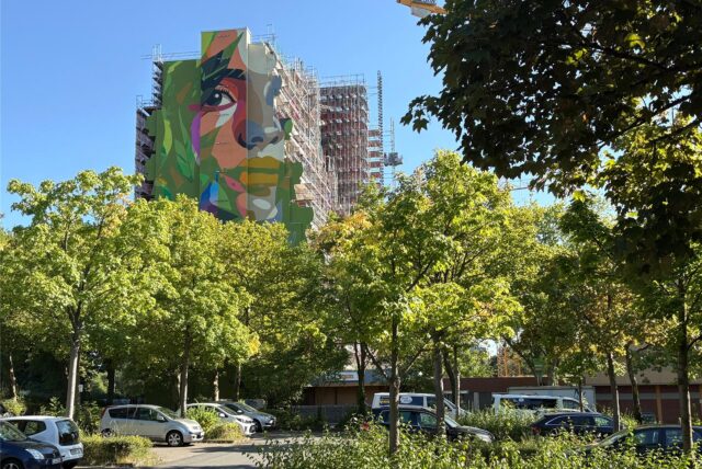 Blick auf die Seite des Hannibals, die mit einem großen Gesicht verziert ist, davor ein Parkplatz mit grünen Bäumen, blauer Himmel
