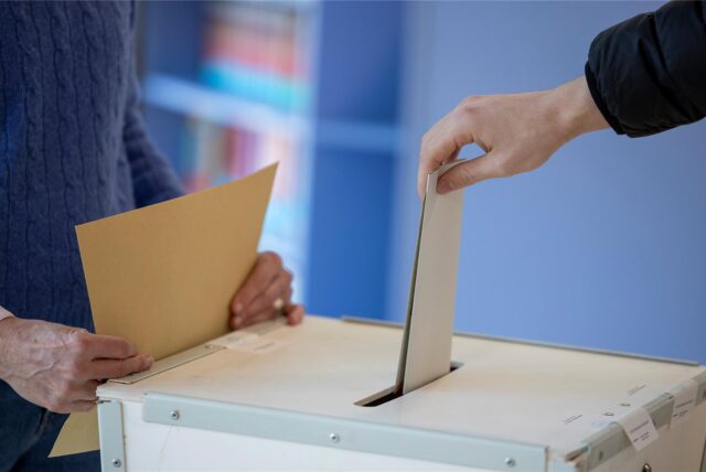 Ein Wähler wirft seinen Stimmzettel in eine Wahlurne. Im Hintergrund hält eine Person einen Umschlag bereit.