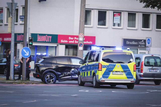 Ein schwarzes Auto mit Dellen in der Seite, davor steht ein Polizeiwagen mit eingeschaltetem Blaulicht