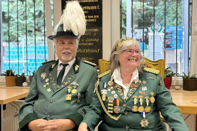 Zwei Personen in grünen Uniformen mit zahlreichen Auszeichnungen posieren für ein Foto. Der Mann trägt einen Hut mit weißen Federn, während die Frau eine goldene Krone trägt.