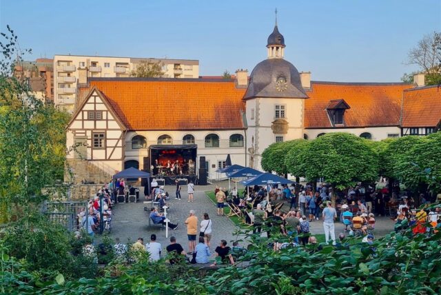 Menschen stehen und sitzen rund um das Wasserschloss Haus Rodenberg in Dortmund-Aplerbeck: Rock am Schloss 2024.