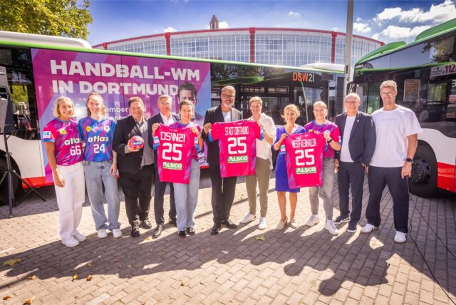 Eine Gruppe von Personen posiert vor einem Bus mit der Aufschrift „Handball-WM in Dortmund“ und hält Trikots mit der Nummer 25. Im Hintergrund ist die Westfalenhalle zu sehen.