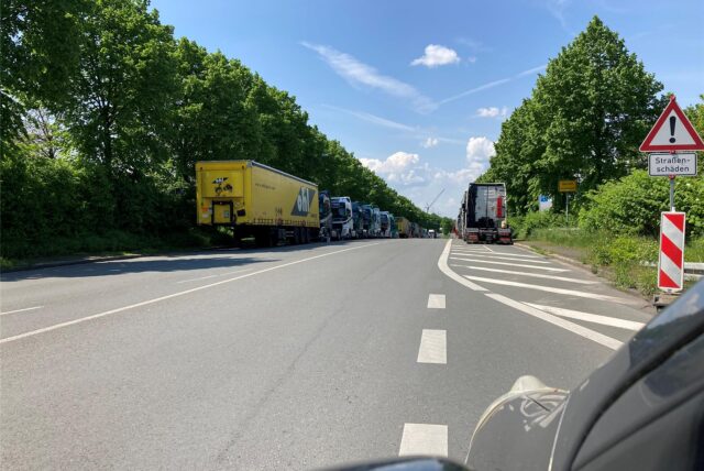 Auf einer Landstraße in der Nähe von Herten staut sich der Verkehr, bestehend aus mehreren Lastkraftwagen. Ein Warnschild weist auf Straßenschäden hin.