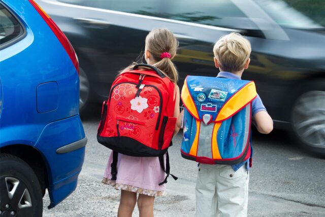 Zwei Schulkinder stehen am Straßenrand neben einem geparkten Pkw.